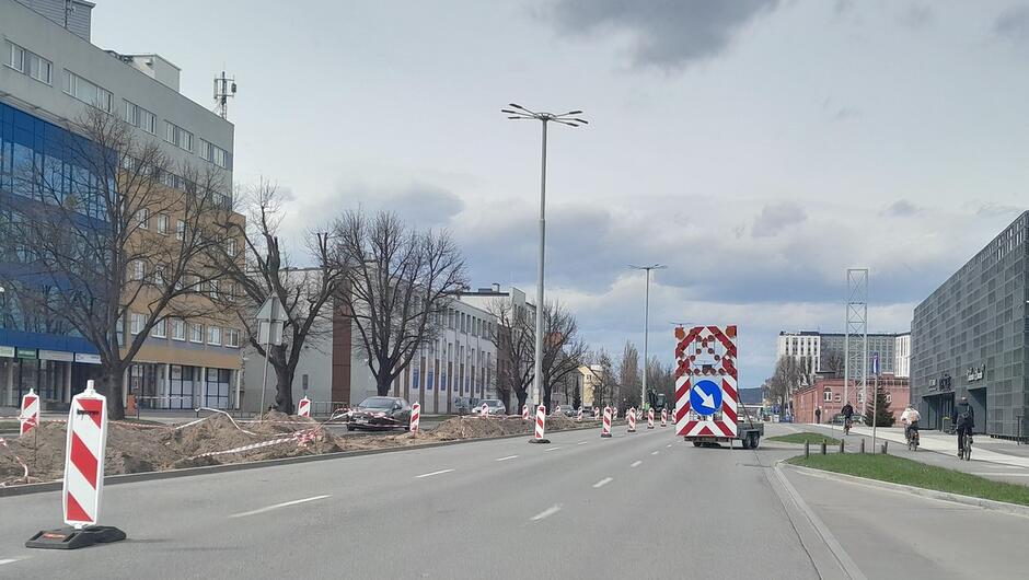 Zdjęcie przedstawia odcinek miejskiej ulicy objęty robotami drogowymi. Ruch pojazdów jest częściowo ograniczony i odpowiednio oznakowany za pomocą przenośnych znaków oraz zapór drogowych. Na jezdni ustawiona jest przyczepa z tablicą ostrzegawczą, informująca o konieczności zmiany toru jazdy. Wzdłuż drogi widoczne są wykopy zabezpieczone taśmami ostrzegawczymi. W tle znajdują się budynki użyteczności publicznej i biurowe.