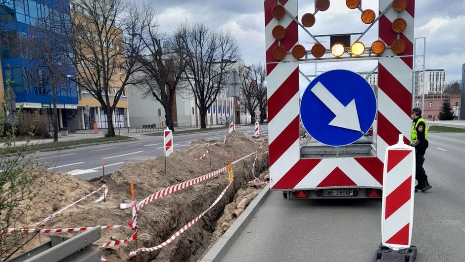 Na fotografii widoczna jest przyczepa drogowa z tablicą świetlną i znakiem nakazującym ominięcie przeszkody z prawej strony. Obok znajduje się funkcjonariusz (prawdopodobnie policji lub służb porządkowych), który nadzoruje organizację ruchu. Po lewej stronie widoczny jest głęboki wykop w pasie drogowym, zabezpieczony pachołkami i taśmami. Całość wskazuje na aktywnie prowadzone prace drogowe, wymagające szczególnej ostrożności uczestników ruchu.