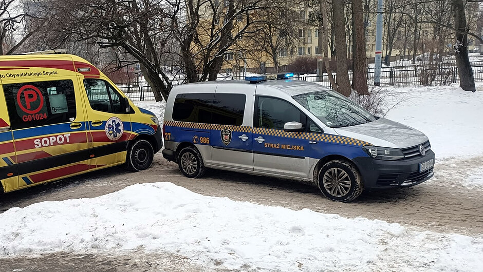 Radiowóz straży miejskiej oraz ambulans stoją na zaśnieżonej drodze w rejonie osiedla mieszkaniowego. Na miejsce interwencji przyjechali strażnicy miejscy oraz ratownicy medyczni, którzy wspólnie podjęli działania wobec starszej kobiety wymagającej pomocy. Fotografia dokumentuje współpracę służb w czasie zimowych warunków.