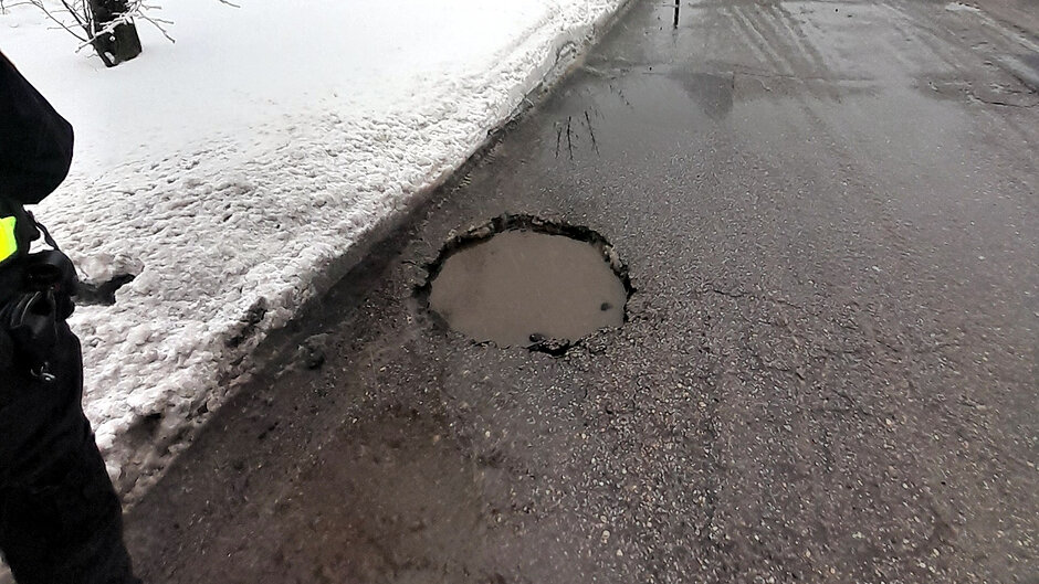 Na jezdni widoczny jest sporych rozmiarów ubytek w asfalcie wypełniony wodą, znajdujący się tuż przy krawężniku. Dziura stanowi potencjalne zagrożenie dla kierowców. Fotografia została wykonana w zimowych warunkach, przy zalegającym śniegu, podczas interwencji strażników miejskich.