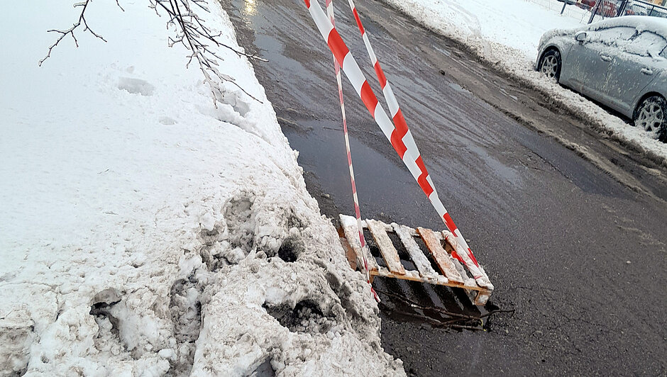 Zabezpieczony ubytek w jezdni oznaczony paletą oraz taśmą ostrzegawczą. Niebezpieczne miejsce znajduje się przy krawężniku, w pobliżu zaśnieżonego pobocza. Oznakowanie ma na celu ostrzeżenie kierowców przed zagrożeniem do czasu przyjazdu służb drogowych.