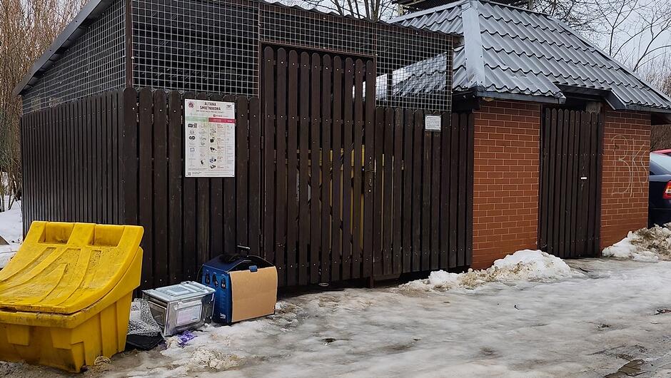 Widzimy zimową scenę przedstawiającą altanę śmietnikową na tle osiedla. Konstrukcja składa się z ciemnych, drewnianych pionowych desek oraz fragmentów czerwonej cegły, a całość przykryta jest szarym, metalowym dachem. Wejście zabezpiecza brama z metalowej siatki, na której umieszczono tablicę informacyjną z napisem „ALTANA ŚMIETNIKOWA” oraz zasadami segregacji odpadów w języku polskim. Przed altaną stoją różne przedmioty pozostawione na śniegu: duży żółty plastikowy pojemnik, niebiesko‑czarne urządzenie lub sprzęt, kartonowe pudełko oraz metalowy kosz. Wokół zalega śnieg i lód, co podkreśla zimową aurę. Po prawej stronie kadru widoczny jest fragment zaparkowanego samochodu. Całość sprawia wrażenie typowego, uporządkowanego miejsca do gromadzenia odpadów na terenie osiedla mieszkaniowego.