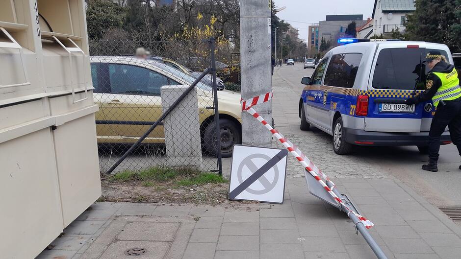 Na zdjęciu widoczny jest fragment chodnika oraz pasa drogowego w obszarze zabudowanym. Na pierwszym planie znajduje się złamany metalowy słupek ze znakiem drogowym, który został wyrwany z podłoża i opiera się o betonowy słup. Znak jest przechylony i zabezpieczony biało‑czerwoną taśmą ostrzegawczą, rozciągniętą pomiędzy słupkiem a elementem infrastruktury. W tle, za ogrodzeniem z siatki, stoi samochód osobowy koloru złotego, zaparkowany w pobliżu posesji. Po prawej stronie widoczny jest radiowóz policyjny z włączonymi niebieskimi światłami sygnalizacyjnymi oraz osoba wykonująca czynności przy pojeździe. Nawierzchnia chodnika wykonana jest z płyt betonowych, a okolica ma charakter miejskiej ulicy.