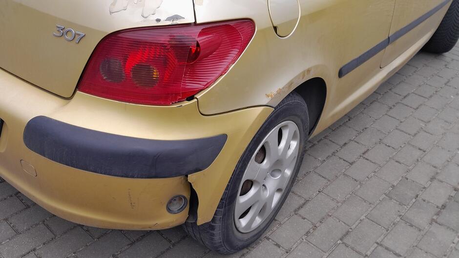 Widoczne są uszkodzenia tylnego zderzaka, w tym odkształcenia, rysy oraz miejscowe pęknięcia w dolnej części. Na błotniku w pobliżu nadkola zauważalne są ślady korozji oraz obtarcia lakieru. Tylna lampa znajduje się na swoim miejscu, bez widocznych pęknięć. Samochód stoi na utwardzonej nawierzchni z kostki brukowej.