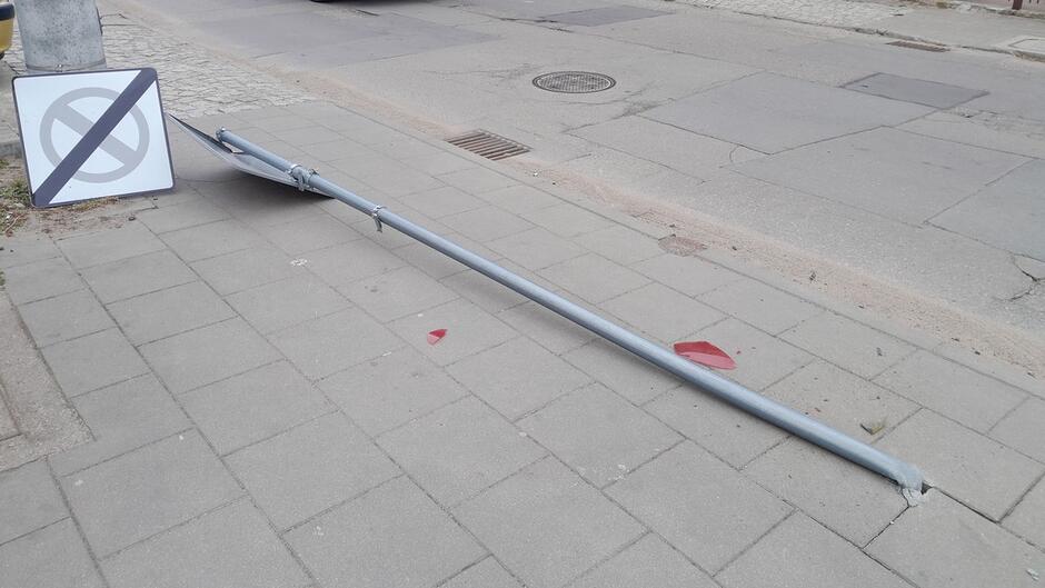 Na zdjęciu widoczny jest metalowy słup drogowy leżący wzdłuż chodnika, całkowicie przewrócony. Na jednym z jego końców znajduje się znak drogowy, który jest wygięty i częściowo uniesiony nad ziemią. Na nawierzchni chodnika, w pobliżu słupa, znajdują się czerwone fragmenty tworzywa, prawdopodobnie elementy rozbitego klosza lub znaku. Chodnik wykonany jest z betonowych płyt, a obok znajduje się jezdnia asfaltowa.