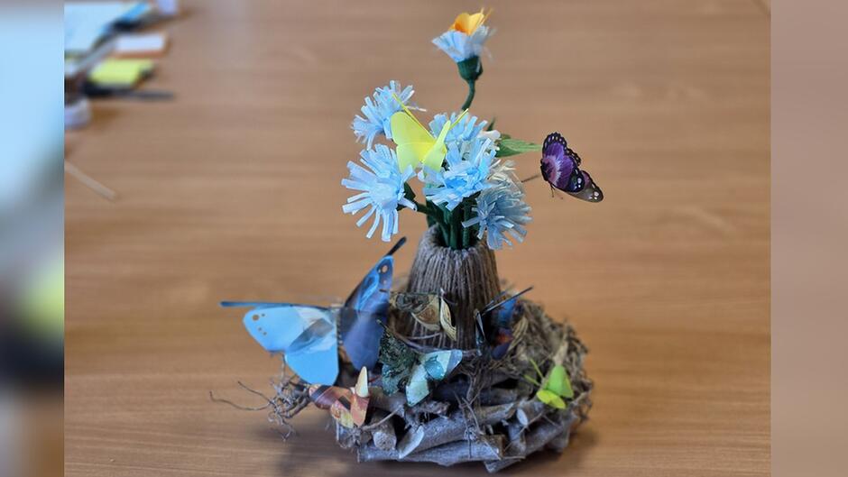 Na pierwszym zdjęciu widoczne są dwie osoby siedzące przy stole, ubrane w ciemne, jednolite koszulki z naszywkami umieszczonymi na rękawach oraz na piersi. Obie osoby mają przypięte niewielkie urządzenia wyglądające jak mikrofony.
Osoby wykonują pracę manualną – jedna z nich trzyma w dłoniach jasnoniebieskie elementy przypominające wstążki lub paski papieru, druga używa nożyczek, wycinając zielony materiał. Na stole leżą prostokątne, jasne podkładki, na których odbywa się praca.
Tło stanowi jasna, drewniana ściana, ozdobiona kolorowymi motylami przyklejonymi na powierzchni. Motyle mają różne kolory, m.in. niebieski, zielony, fioletowy i czerwony, co nadaje przestrzeni dekoracyjny charakter. Całość sprawia wrażenie zajęć plastycznych lub przygotowywania elementów ozdobnych.