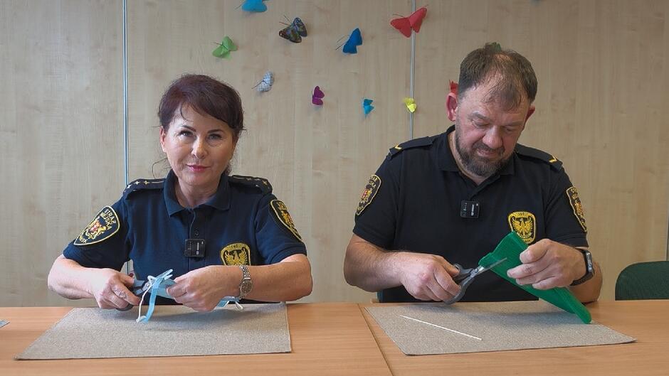 Drugie zdjęcie przedstawia niewielką dekorację ustawioną na drewnianym stole. W centrum znajduje się mały wazonik o naturalnej, brązowej fakturze, przypominającej sznurek lub włókno. W wazoniku umieszczone są sztuczne kwiaty – dominują jasnoniebieskie, drobne kwiaty, a także jeden mniejszy kwiat w kolorach żółtym i białym.
U podstawy wazonika znajduje się okrągła kompozycja z naturalnych materiałów, takich jak suche gałązki, liście i drobne elementy dekoracyjne. Całość ozdobiona jest kolorowymi, dekoracyjnymi motylami, które różnią się barwą i wielkością – widoczne są m.in. motyle w odcieniach niebieskiego, fioletu i zieleni.
Kompozycja sprawia wrażenie ręcznie wykonanego stroika dekoracyjnego, inspirowanego motywami przyrody i wiosny.