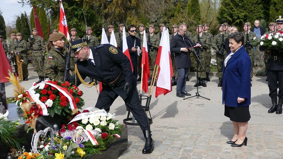 Strażnik miejski składa kwiaty pod pomnikiem Golgota Wschodu, za nim stoi przedstawicielka Wojewody Pomorskiej.