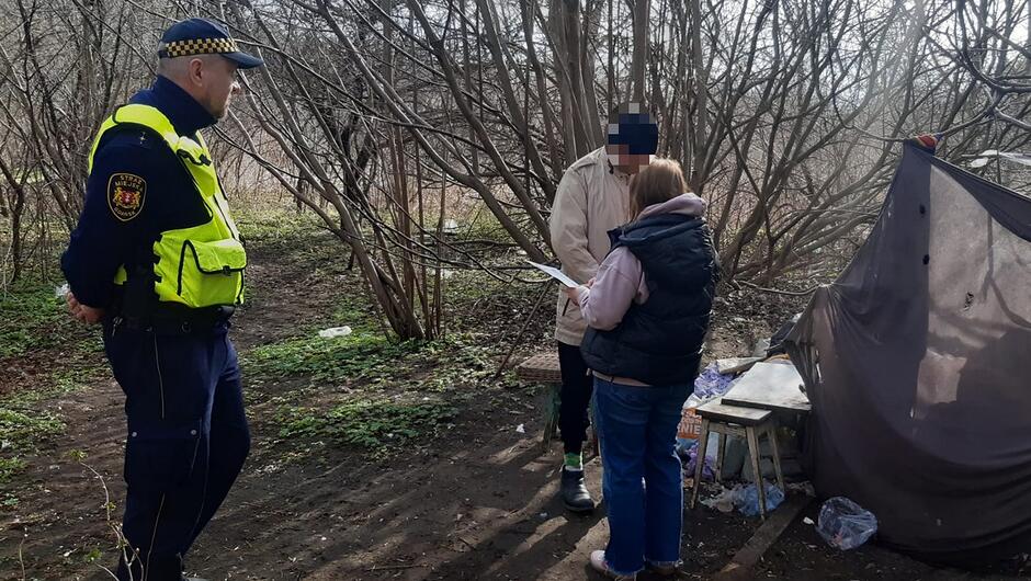 Na zdjęciu widać strażnika miejskiego w żółtej kamizelce odblaskowej, który stoi w lesistym terenie obok dwóch innych osób. Strażnik prowadzi z nimi interwencję lub kontrolę przy prowizorycznym obozowisku zbudowanym z ciemnego materiału. Obok schronienia leżą różne rzeczy osobiste, co sugeruje, że to miejsce tymczasowego pobytu. Jedna z osób trzyma dokumenty, co wygląda na formalną rozmowę z funkcjonariuszem.