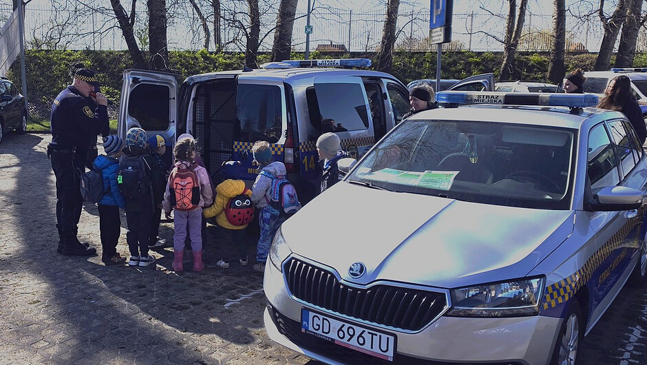 Zdjęcie wykonane na zewnątrz, na utwardzonym placu lub parkingu. Na pierwszym planie widoczny jest oznakowany pojazd służbowy z niebiesko-żółtymi elementami graficznymi i tablicą rejestracyjną. Obok stoi drugi, podobnie oznakowany samochód z otwartymi tylnymi drzwiami.
Pomiędzy pojazdami zgromadzona jest grupa małych dzieci w kolorowych kurtkach i plecakach. Obok grupy stoją osoby dorosłe w umundurowaniu. W tle widoczne są drzewa bez liści, ogrodzenie oraz znaki parkingowe, co sugeruje teren miejski. Warunki pogodowe wskazują na dzień słoneczny.