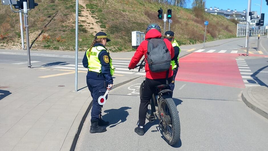 Na drodze pieszo-rowerowej strażnik miejski prowadzi czynności wobec rowerzysty. Obok zaparkowany jest oznakowany pojazd Straży Miejskiej. Kontrola dotyczy zasad poruszania się rowerem w przestrzeni publicznej.