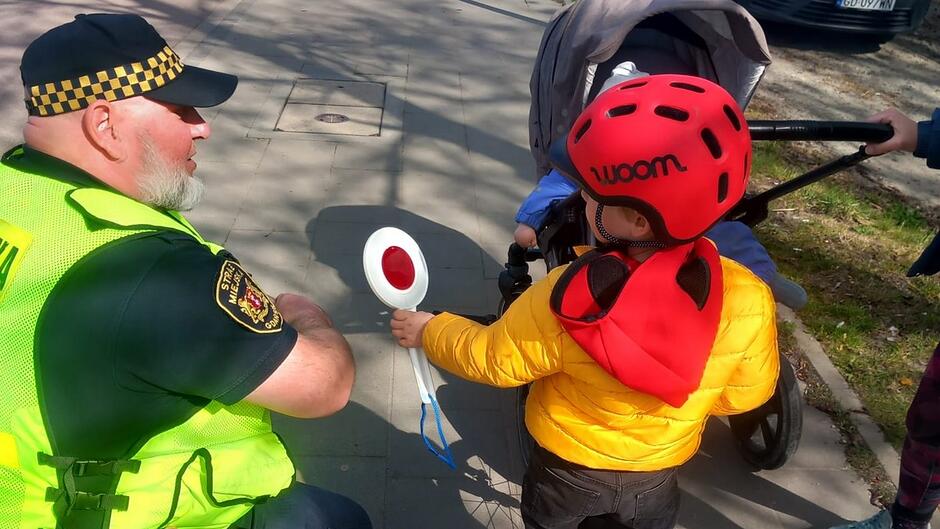 Strażnik miejski prowadzi rozmowę z dzieckiem poruszającym się na rowerze pod opieką osoby dorosłej. Działania mają charakter edukacyjny i dotyczą zasad bezpiecznego korzystania z roweru w przestrzeni miejskiej.