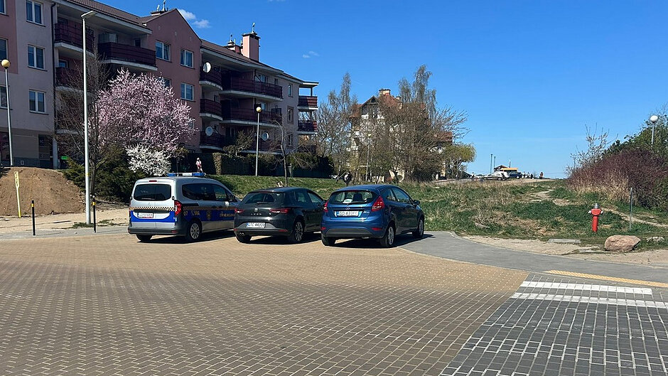 Zdjęcie przedstawia parking z nawierzchnią wykonaną z kostki brukowej. Zaparkowane są trzy samochody, w tym jeden pojazd służbowy. W tle widoczna jest zabudowa mieszkaniowa – kilkukondygnacyjne budynki oraz tereny zielone z krzewami i drzewami. Po prawej stronie znajduje się hydrant przeciwpożarowy. Cała scena jest dobrze oświetlona światłem dziennym, przy bezchmurnym niebie.