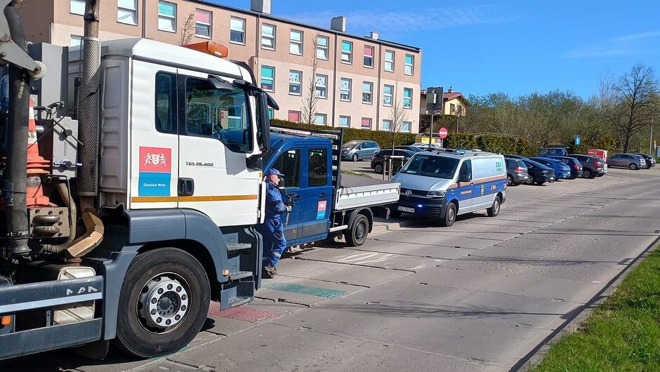 Zdjęcie przedstawia ulicę na obszarze zabudowy mieszkaniowej. Po lewej stronie widoczny jest duży pojazd specjalistyczny z oznaczeniami „Gdańskie Wody”, zaparkowany przy krawędzi jezdni. Obok znajduje się mniejszy samochód dostawczy oraz pojazd służbowy z oznaczeniami miejskimi. W tle znajdują się zaparkowane samochody osobowe oraz kilkukondygnacyjny budynek mieszkalny. Warunki pogodowe są dobre, z wyraźnym, słonecznym oświetleniem.