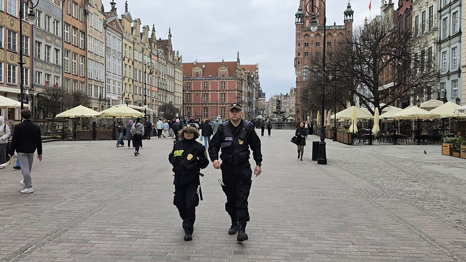 Dwie osoby w mundurach Straży Miejskiej idą szerokim, brukowanym placem (Długim Targiem) w historycznej części Gdańska. Po obu stronach placu stoją kolorowe kamienice.