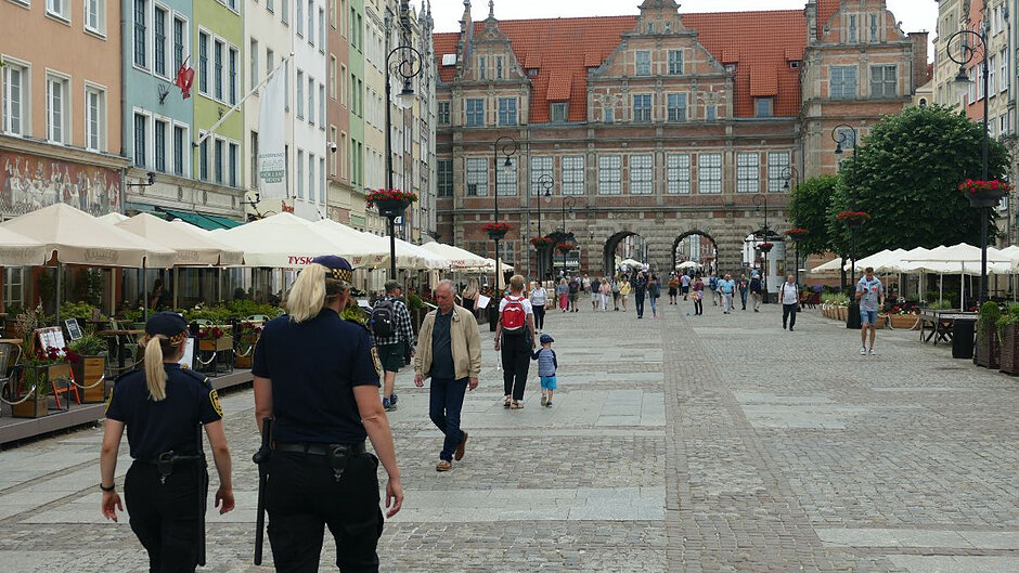 Dwie strażniczki miejskie idą szerokim, wybrukowanym placem (Długim Targiem). Po obu stronach kolorowe kamieniczki, a także ogródki kawiarniane z parasolami. 