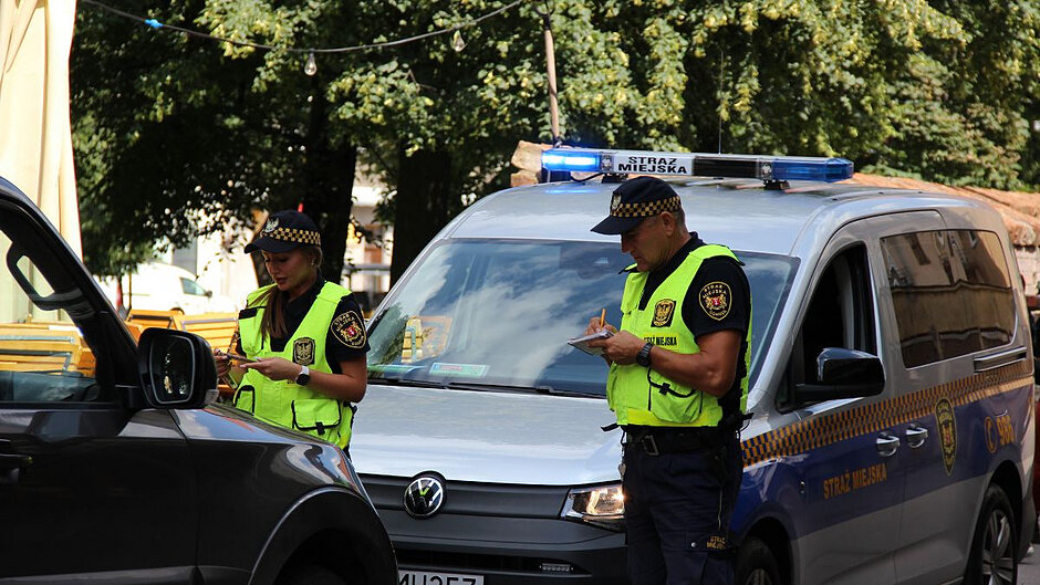 Strażniczka i strażnik w kamizelkach odblaskowych stoją pomiędzy radiowozem a samochodem osobowym i coś notują. 