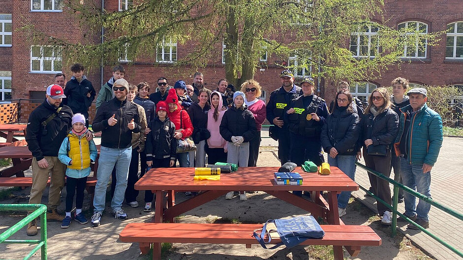 Zdjęcie przedstawia trzy osoby stojące przy ogrodzeniu na skraju terenu zielonego. Dwie z nich mają na sobie umundurowanie z elementami odblaskowymi i oznaczeniem „STRAŻ MIEJSKA”. Przed nimi na ziemi ułożony jest stos worków w kolorach żółtym, zielonym i czarnym, wypełnionych zebranymi odpadami. W tle widoczne są drzewa i krzewy, część z nich kwitnąca, co sugeruje wiosenną porę roku. Scena dokumentuje zakończoną akcję porządkową w terenie.