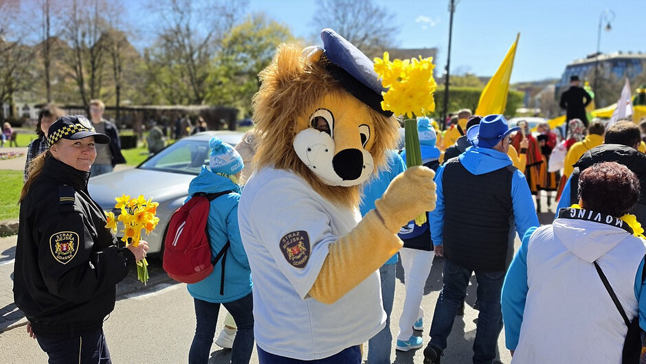 Fotografia z wydarzenia plenerowego na ulicy lub w parku miejskim. Na pierwszym planie widoczna jest maskotka w stroju zwierzęcia, trzymająca bukiet żółtych kwiatów oraz żółtą flagę. Obok znajduje się osoba w umundurowaniu z naszywką „STRAŻ MIEJSKA GDAŃSK”. W tle widoczna jest większa grupa uczestników ubranych w niebieskie i jasne kurtki, również niosących żółte elementy. Otoczenie stanowi zieleń miejska, zabudowania oraz zaparkowane samochody.