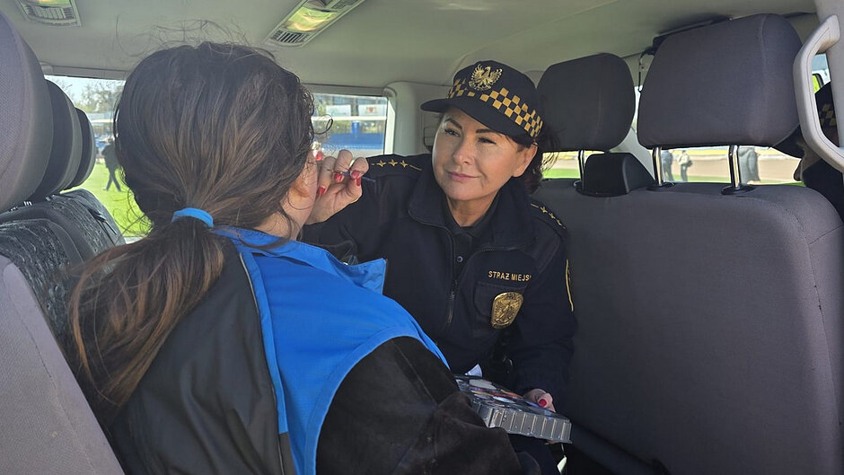 Zdjęcie wykonane wewnątrz pojazdu. Osoba w umundurowaniu z wyraźnym napisem „STRAŻ MIEJSKA” na iemblemacie siedzi na tylnej kanapie i wykonuje makijaż innej osoby siedzącej przodem. Widoczne są pędzelki oraz paleta kosmetyków. Przez okna pojazdu można dostrzec teren zielony i fragment infrastruktury sportowej. Scena ma charakter przygotowań do występu lub wydarzenia.