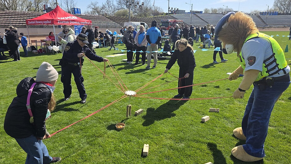 Fotografia wykonana na otwartym stadionie lub boisku sportowym. Kilkoro uczestników stoi w okręgu na trawie i trzyma długie czerwone linki przymocowane do drewnianej konstrukcji znajdującej się na środku. Konstrukcja otoczona jest niewielkimi klockami i misą. W tle widać namioty informacyjne, osoby obserwujące wydarzenie oraz trybuny stadionowe. Jedna z osób ma na sobie elementy umundurowania, inna kostium maskotki. Wygląda to na aktywność integracyjną lub edukacyjną w ramach wydarzenia plenerowego.