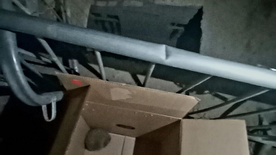 Fotografia wykonana jest w ciemnym, zamkniętym miejscu, przypominającym piwnicę lub przestrzeń techniczną. W dolnej części kadru znajduje się otwarta kartonowa skrzynka, w której widoczny jest zwinięty, jasnoszary obiekt (może być to element materiału lub zwierzę zwinięte w kłębek – brak jednoznacznych detali). Wokół skrzynki biegną metalowe rury i instalacje. Ściany są surowe, betonowe, a oświetlenie jest słabe i punktowe, co powoduje wyraźne cienie.