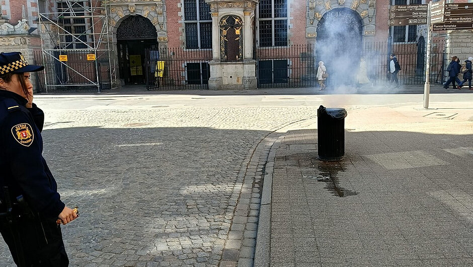 Fotografia przedstawia fragment ulicy z zabytkową zabudową w tle. Widoczna jest ceglana elewacja budynku z licznymi oknami oraz ozdobnymi detalami architektonicznymi, w tym niszami z rzeźbami i portalami. Po lewej stronie zdjęcia znajduje się konstrukcja rusztowania. Na pierwszym planie widać osobę w umundurowaniu, stojącą na brukowanej nawierzchni. W centralnej części kadru unosi się biały dym lub para, wydobywająca się z czarnego kosza ulicznego. W tle poruszają się przechodnie. Całość wykonana jest w świetle dziennym.