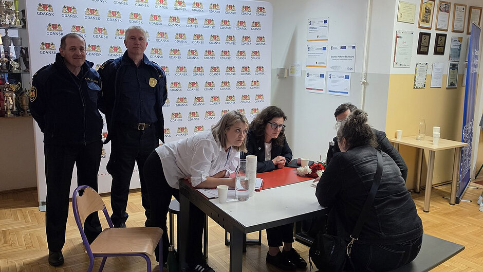Uczestnicy spotkania „Porozmawiajmy przy stole” prowadzą bezpośrednią rozmowę z mieszkańcem. W tle widoczni są strażnicy miejscy oraz ścianka z herbem Gdańska.