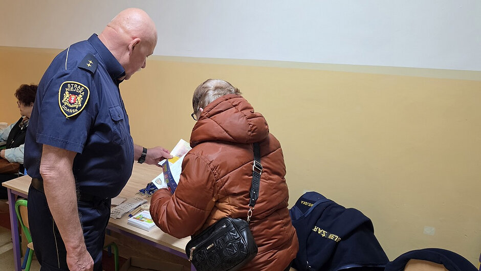 Strażnik miejski udziela informacji mieszkańcowi przy stoisku informacyjnym i przekazuje materiały edukacyjne dotyczące bezpieczeństwa.