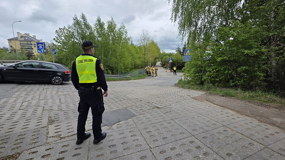 Na pierwszym planie stoi funkcjonariusz Straży Miejskiej w czarnym mundurze i żółtej kamizelce odblaskowej z napisem STRAŻ MIEJSKA na plecach. Patrzy w kierunku ścieżki prowadzącej w głąb zalesionego terenu. Po lewej stronie przy krawędzi drogi zaparkowany jest czarny samochód. Tło stanowi zielony, gęsty las, a niebo jest zachmurzone.

Dalej, w głębi kadru, widać kilku strażaków w jasnych ubraniach bojowych i kaskach. Stoją przy wejściu na wąską ścieżkę lub mostek, jakby prowadzili działania operacyjne lub sprawdzali teren. Obok nich znajdują się niebieskie znaki informacyjne.

Całość wygląda na interwencję służb miejskich i straży pożarnej w spokojnej, leśnej okolicy.