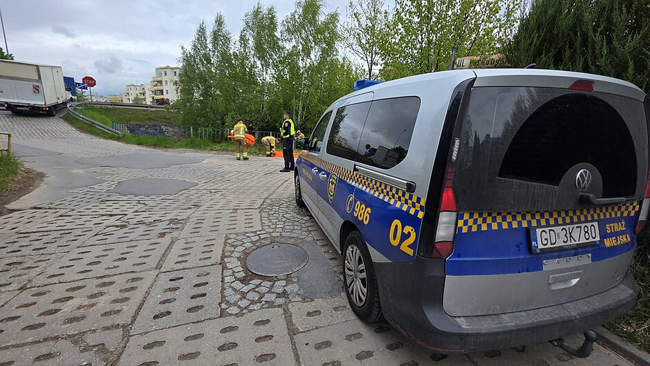 Na zdjęciu przedstawiona jest scena działań służb miejskich na utwardzonej, lokalnej drodze o nawierzchni z płyt betonowych i fragmentów asfaltu, która prowadzi w kierunku niewielkiego wzniesienia.
Otoczenie i tło
W tle widoczne są drzewa i krzewy tworzące zielony pas wzdłuż drogi. Dalej, po lewej stronie, znajdują się jasne budynki mieszkalne o kilku kondygnacjach. Na końcu drogi widoczny jest znak „STOP” ustawiony przy skrzyżowaniu. Niebo jest zachmurzone, co nadaje scenie stonowane, lekko szare oświetlenie.
Pierwszy plan
Po prawej stronie kadru stoi oznakowany samochód Straży Miejskiej marki Volkswagen Caddy. Na pojeździe widoczne są napisy „STRAŻ MIEJSKA”, numer jednostki „986” oraz numer boczny „02”. Samochód ma zamontowane niebieskie światła ostrzegawcze.
Działania służb
W środkowej części zdjęcia znajdują się funkcjonariusz Straży Miejskiej oraz dwie osoby w odzieży ochronnej, prawdopodobnie strażacy. Osoby te pracują przy rozkładaniu długiej, pomarańczowej zapory lub rękawa, który leży na drodze i ciągnie się w stronę barierek oraz zieleni. Jedna z osób pochyla się nad materiałem, wykonując czynności zabezpieczające.
Szczegóły dodatkowe Po lewej stronie widoczny jest duży pojazd ciężarowy stojący przy wzniesieniu drogi.
Droga posiada liczne nierówności i pęknięcia, co jest szczególnie widoczne w dolnej części kadru.
Wzdłuż drogi biegną metalowe barierki oddzielające jezdnię od terenu zielonego.

Wrażenie ogólne
Całość przedstawia zorganizowaną akcję zabezpieczającą prowadzoną przez służby, prawdopodobnie związaną z ochroną terenu lub reagowaniem na potencjalne zagrożenie.