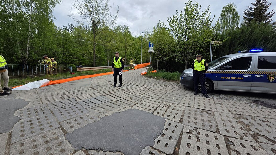 Zdjęcie przedstawia miejsce prowadzenia działań służb na krętej, lokalnej drodze o nawierzchni z betonowych płyt z charakterystycznymi otworami oraz fragmentów popękanego asfaltu.
Otoczenie i tło
Scena rozgrywa się w otoczeniu gęstej zieleni – wzdłuż drogi rosną drzewa i krzewy, a po lewej stronie znajduje się metalowa barierka oddzielająca jezdnię od terenu zielonego. W tle widoczny jest znak drogowy wskazujący kierunek jazdy. Niebo jest pochmurne, co nadaje zdjęciu stonowane, chłodne oświetlenie.
Pierwszy plan i pojazd
Po prawej stronie zaparkowany jest oznakowany samochód Straży Miejskiej z włączonym niebieskim światłem ostrzegawczym. Na pojeździe widoczny jest napis „STRAŻ MIEJSKA”. Obok pojazdu stoi funkcjonariusz w kamizelce odblaskowej.
Działania służb
Na środku drogi oraz po lewej stronie znajdują się kolejne osoby w odzieży roboczej i ochronnej, które rozciągają długą, intensywnie pomarańczową zaporę lub rękaw. Materiał ten biegnie wzdłuż jezdni i w kierunku barierek, co sugeruje zabezpieczanie terenu lub kontrolowanie przepływu substancji.
Jedna z osób stoi na środku drogi, obserwując sytuację, natomiast inne zajmują się układaniem i rozwijaniem zapory.
Dodatkowe szczegóły
Na jezdni, w pobliżu miejsca pracy służb, leży także biały materiał lub element wyposażenia. Nawierzchnia drogi jest wyraźnie zużyta, z licznymi pęknięciami i nierównościami.
Wrażenie ogólne
Zdjęcie dokumentuje trwającą akcję zabezpieczającą prowadzoną przez Straż Miejską oraz inne służby. Teren jest kontrolowany, a działania koncentrują się na rozstawieniu pomarańczowej zapory wzdłuż drogi.