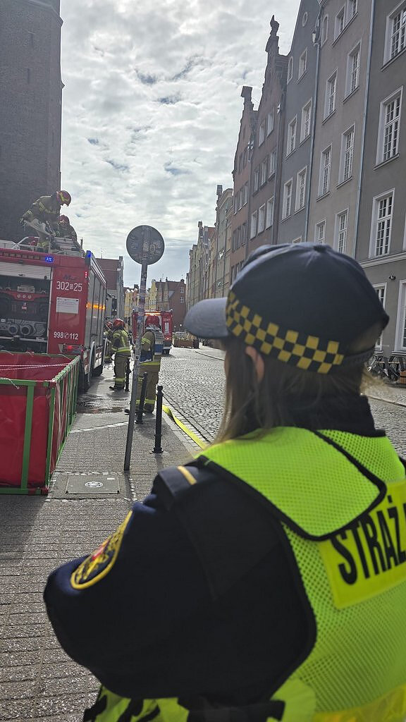 Zdjęcie przedstawia działania służb ratunkowych i porządkowych na ulicy w zabytkowej części miasta.
Otoczenie i tło
Scena rozgrywa się na wąskiej, brukowanej ulicy otoczonej wysokimi kamienicami o ozdobnych fasadach. Po lewej stronie widoczny jest masywny, ceglasty budynek, natomiast po prawej ciąg jasnych kamienic z licznymi oknami. Na końcu ulicy widać kolejne zabudowania, a nad całością rozciąga się zachmurzone niebo z prześwitującym światłem słonecznym.
Pierwszy plan
Na pierwszym planie znajduje się funkcjonariusz Straży Miejskiej stojący tyłem do aparatu. Osoba ta ubrana jest w ciemny mundur oraz jaskrawą kamizelkę odblaskową z napisem „STRAŻ MIEJSKA”. Na głowie ma czapkę z charakterystycznym żółtym wzorem szachownicy.
Działania służb
W środkowej części zdjęcia widać kilka osób w odzieży ochronnej, prawdopodobnie strażaków, którzy pracują przy wozie strażackim zaparkowanym przy krawędzi ulicy. Jeden z ratowników znajduje się na górze pojazdu, obsługując sprzęt, a pozostali stoją obok i wykonują czynności związane z akcją.
Obok wozu ustawiony jest czerwony, mobilny zbiornik lub zapora w metalowej konstrukcji, podobny do tymczasowego basenu, który może służyć do gromadzenia wody lub innych substancji. Element ten jest rozstawiony przy chodniku, oddzielony od jezdni niewielkimi słupkami.
Szczegóły dodatkowe Na jezdni widoczny jest znak drogowy zamontowany na słupie.
W pobliżu znajduje się drugi pojazd służb w tle.
Nawierzchnia ulicy jest wykonana z kostki brukowej, charakterystycznej dla starego miasta.
Cały teren wygląda na zabezpieczony i kontrolowany przez służby.

Wrażenie ogólne
Zdjęcie ukazuje skoordynowaną akcję Straży Pożarnej oraz Straży Miejskiej w przestrzeni miejskiej. Funkcjonariusz na pierwszym planie obserwuje przebieg działań, podczas gdy strażacy obsługują sprzęt i przygotowują zabezpieczenia przy użyciu specjalistycznego wyposażenia.