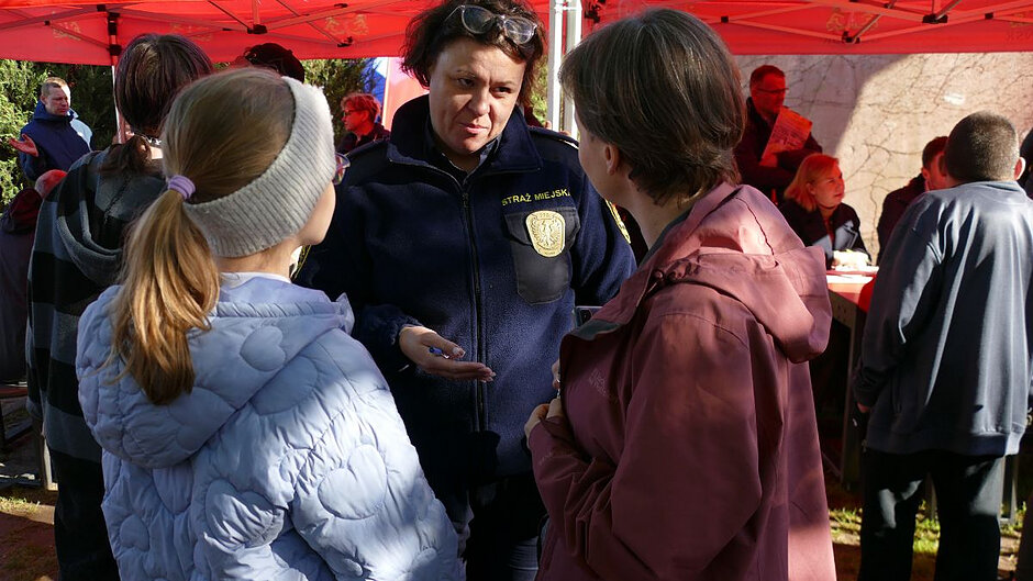 Na zdjęciu widać kilka osób stojących blisko siebie pod czerwonym namiotem plenerowym. W centrum znajduje się strażniczka miejska. Osoba ta prowadzi rozmowę z dwiema innymi osobami stojącymi naprzeciwko, ubranymi w kurtki: jedną jasną pikowaną z opaską na głowie oraz drugą w ciemnoróżowym odcieniu.
W tle znajdują się kolejne osoby oraz stoliki, przy których siedzą uczestnicy wydarzenia. 