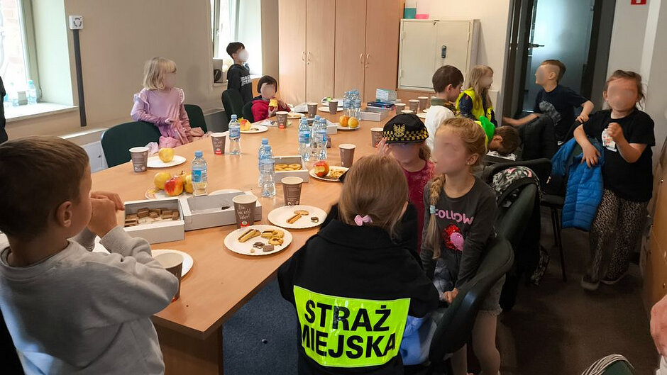 Na zdjęciu przedstawiono wnętrze jasnego pomieszczenia biurowego lub świetlicy. W centralnej części znajduje się duży, prostokątny stół z jasnego drewna, wokół którego siedzi i stoi grupa dzieci. Na stole ustawiono jednorazowe kubki, talerzyki z jedzeniem (m.in. kawałki owoców, słodycze, kanapki lub przekąski) oraz kilka butelek wody. Widoczne są również pudełka, prawdopodobnie po przekąskach.
Wokół stołu znajduje się kilkanaście dzieci, niektóre siedzą na krzesłach, inne stoją lub przemieszczają się w pomieszczeniu. W tle, przy ścianach, stoją szafki i regały, a także zamknięta skrzynka lub szafa na dokumenty. W jednym z narożników widać drzwi.
Na pierwszym planie, tyłem do aparatu, siedzi osoba w ciemnym stroju z wyraźnym napisem „STRAŻ MIEJSKA” w kolorze jaskrawym na plecach, co sugeruje obecność przedstawiciela służb miejskich. Obok tej osoby znajduje się dziecko w czapce przypominającej element stroju służb mundurowych.
Oświetlenie jest naturalne i jasne, prawdopodobnie z okna znajdującego się po lewej stronie. Całość sprawia wrażenie spotkania edukacyjnego lub integracyjnego, podczas którego dzieci spożywają posiłek i przebywają w towarzystwie osoby reprezentującej straż miejską.