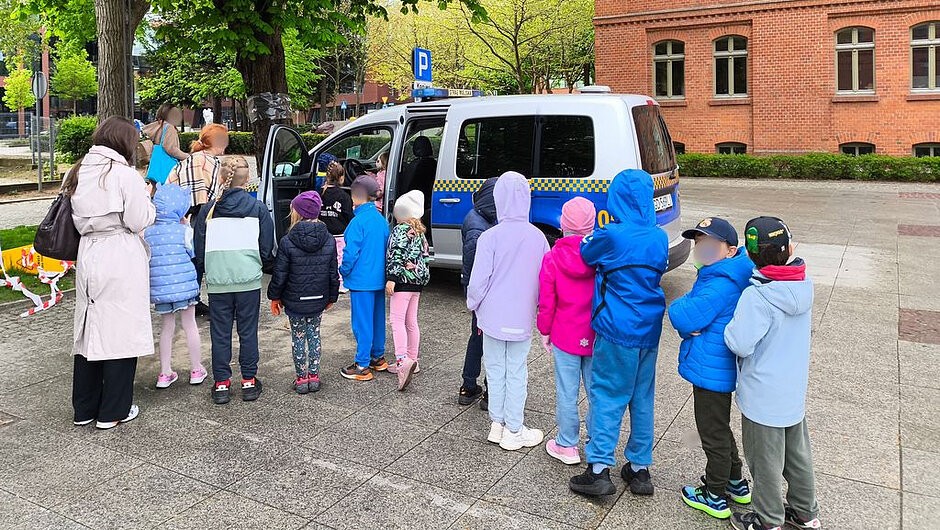 Na zdjęciu widać scenę na zewnątrz, na utwardzonym placu lub chodniku, otoczonym zielenią oraz budynkami z czerwonej cegły. W centrum znajduje się oznakowany samochód służbowy (biało-niebieski van z charakterystycznym pasem i światłami na dachu). Jedno z bocznych drzwi pojazdu jest otwarte.
Przy samochodzie stoi grupa kilkunastu dzieci ustawionych w kolejce. Dzieci mają na sobie kurtki i czapki, co sugeruje chłodniejszą pogodę. Część dzieci patrzy w stronę wnętrza pojazdu, a niektóre są już przy drzwiach i zaglądają do środka. Obok dzieci znajduje się osoba dorosła w długim płaszczu, stojąca z lewej strony, prawdopodobnie opiekun grupy.
W tle widoczne są drzewa z zielonymi liśćmi, znak parkingowy oraz fragment ulicy. Po prawej stronie znajduje się budynek z czerwonej cegły z oknami. Na chodniku ustawiono także kolorowe elementy oznakowania lub zabaw edukacyjnych.
Całość przedstawia prawdopodobnie zajęcia edukacyjne w plenerze, podczas których dzieci poznają pojazd służbowy i jego wyposażenie.
