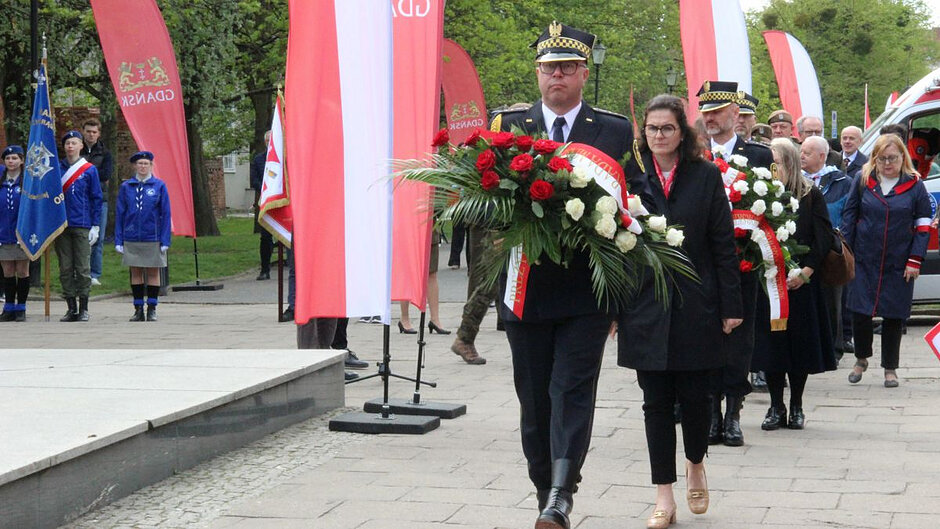 Strażnik niesie wieniec, za nim prezydent Gdańska Aleksandra Dulkiewicz oraz kolejne oficjalne reprezentacje zamierzające złożyć kwiaty pod pomnikiem Tym co za polskość Gdańska . Po lewej flagi Polski i gdańskie oraz w tle widoczna warta trójki harcerzy. 