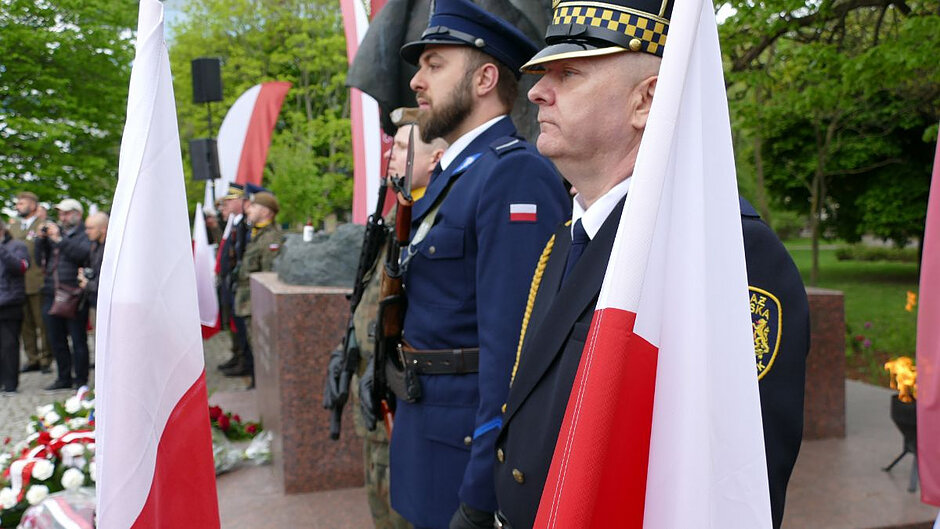 Na pierwszym planie dwie flagi Polski oraz strażnik miejski, policjant i żołnierz pełniący wartę honorową przy pomniku Józefa Piłsudskiego. 