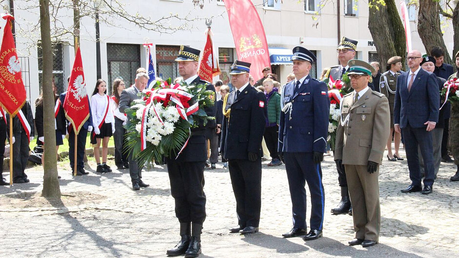 Strażnik miejski stoi, trzymając wieniec, za nim trzej przedstawiciele służb mundurowych. W tle uczniowskie poczty sztandarowe. 