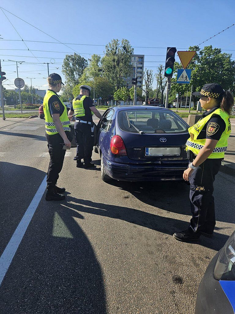 Na zdjęciu przedstawiono interwencję drogową prowadzoną przez funkcjonariuszy Straży Miejskiej na skrzyżowaniu ulic w terenie miejskim.
Na pierwszym planie widoczny jest zatrzymany samochód osobowy marki Toyota w kolorze ciemnoniebieskim, z numerem rejestracyjnym „GD 4546H”. Pojazd stoi przy krawędzi pasa ruchu, tuż przed linią zatrzymania. Obok samochodu znajduje się trzech funkcjonariuszy w umundurowaniu – mają na sobie ciemne ubrania służbowe oraz kamizelki odblaskowe z oznaczeniami Straży Miejskiej. Jeden z funkcjonariuszy stoi przy drzwiach kierowcy i prowadzi czynności kontrolne, natomiast pozostali obserwują sytuację z boku.
W tle widoczne jest skrzyżowanie z sygnalizacją świetlną – światło dla pojazdów jest zielone. Obecne są również znaki drogowe, w tym znak „ustąp pierwszeństwa” oraz oznaczenie przejścia dla pieszych. Nad skrzyżowaniem rozciąga się sieć trakcji tramwajowej, co wskazuje na lokalizację w większym mieście. W dalszej części kadru znajdują się drzewa oraz budynki mieszkalne.
Oświetlenie wskazuje na poranne godziny – słońce jest nisko, rzucając długie cienie na jezdnię. Cała scena dokumentuje moment zatrzymania pojazdu i prowadzenia interwencji wobec kierującego.