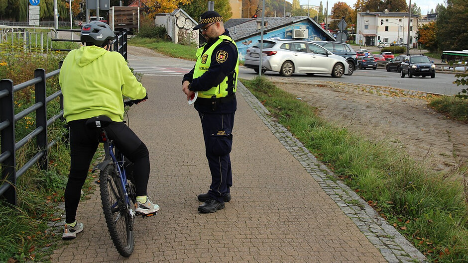 Zdjęcie przedstawia sytuację na chodniku przy ruchliwym skrzyżowaniu w mieście. Widoczny jest fragment infrastruktury drogowej, przejścia dla pieszych oraz ruch samochodowy w tle. Jest słoneczny dzień, a światło jest jasne i naturalne.
Pierwszy plan
Na pierwszym planie znajdują się trzy osoby oraz pojazd Straży Miejskiej:
Dwie osoby w umundurowaniu Straży Miejskiej mają na sobie ciemne stroje oraz jaskrawe, żółto-zielone kamizelki z napisem „STRAŻ MIEJSKA”.
Trzecia osoba stoi naprzeciw funkcjonariuszy – twarz tej osoby jest częściowo rozmyta.
Obok znajduje się biało-niebieski samochód służbowy z napisem „STRAŻ MIEJSKA” oraz numerem rejestracyjnym GD 3G509. Na dachu pojazdu widoczny jest niebieski sygnał świetlny.
Obiekty i ich relacje
Przy krawężniku stoi kilka hulajnóg elektrycznych ustawionych obok siebie.
Po lewej stronie widoczny jest skuter elektryczny.
Dwóch funkcjonariuszy stoi naprzeciwko osoby cywilnej, co sugeruje kontrolę lub rozmowę.
Tło
W tle znajdują się budynki (jeden z nich ma nowoczesną, ciemną elewację), drzewa oraz ruchliwe skrzyżowanie z sygnalizacją świetlną.
Widoczne są inne pojazdy i oznakowanie drogowe, w tym znak przejścia dla pieszych.
Kolory i szczegóły
Dominują kolory: szarość chodnika, zieleń kamizelek, biało-niebieski pojazd oraz naturalne kolory otoczenia. Całość jest dobrze oświetlona.