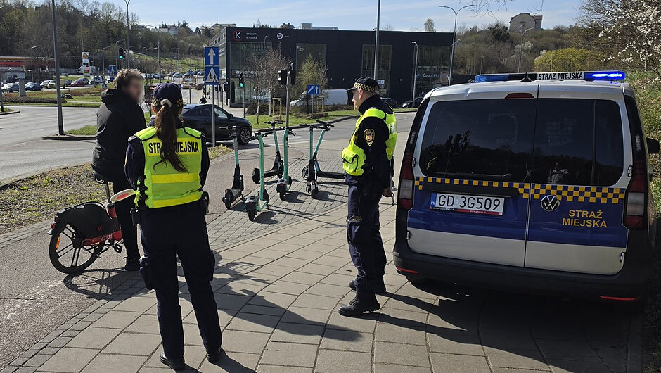 Zdjęcie przedstawia ścieżkę pieszo-rowerową biegnącą wzdłuż ulicy. W tle widoczny jest ruch samochodowy i zabudowa miejska. Otoczenie wskazuje na porę jesienną – na drzewach są kolorowe liście, a część z nich opadła.
Pierwszy plan
Na pierwszym planie znajdują się dwie osoby:
Jedna osoba ubrana jest w jasną, jaskrawą bluzę i stoi przy rowerze górskim, trzymając kierownicę.
Druga osoba to funkcjonariusz Straży Miejskiej w ciemnym mundurze i kamizelce odblaskowej z napisem „STRAŻ MIEJSKA”.
Obiekty i ich relacje
Funkcjonariusz stoi naprzeciw rowerzysty i trzyma w ręku niewielki przedmiot (może to być notatnik lub urządzenie).
Rowerzysta stoi nieruchomo na ścieżce, co sugeruje kontrolę lub rozmowę.
Tło
W tle znajduje się ruch uliczny: kilka samochodów osobowych porusza się po drodze.
Widoczne są ogrodzenia, chodnik, fragmenty trawy oraz budynki mieszkalne i usługowe.
Na poboczu widać także fragment ścieżki gruntowej.
Kolory i szczegóły
Na zdjęciu dominują jesienne barwy: zielenie, żółcie i pomarańcze liści. Ubranie rowerzysty oraz kamizelka funkcjonariusza są jaskrawe, co mocno kontrastuje z otoczeniem.