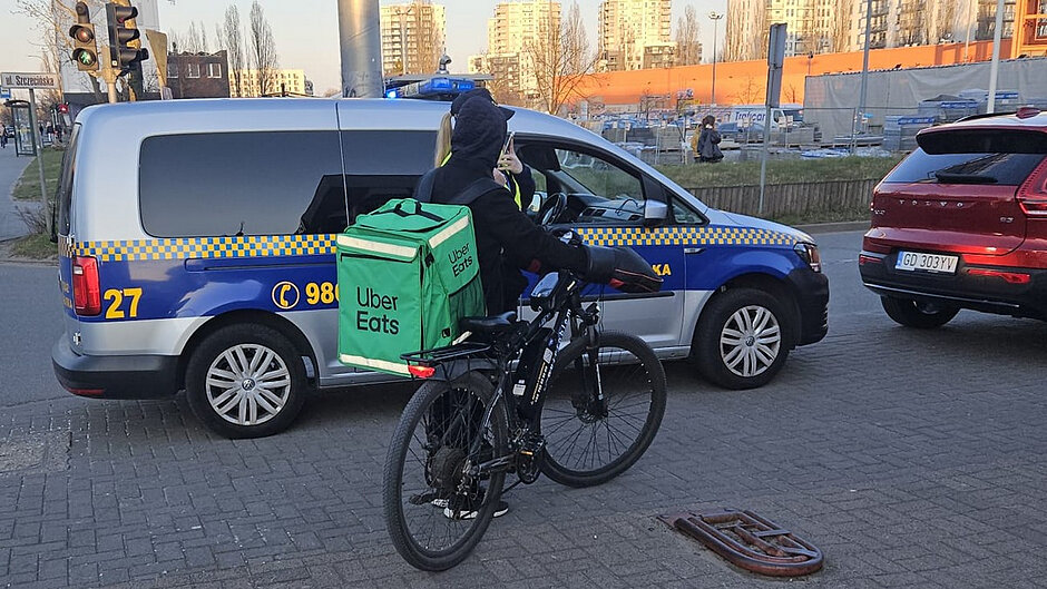Zdjęcie przedstawia ścieżkę pieszo-rowerową biegnącą wzdłuż ulicy. W tle widoczny jest ruch samochodowy i zabudowa miejska. Otoczenie wskazuje na porę jesienną – na drzewach są kolorowe liście, a część z nich opadła.
Pierwszy plan
Na pierwszym planie znajdują się dwie osoby: Jedna osoba ubrana jest w jasną, jaskrawą bluzę i stoi przy rowerze górskim, trzymając kierownicę.
Druga osoba to funkcjonariusz Straży Miejskiej w ciemnym mundurze i kamizelce odblaskowej z napisem „STRAŻ MIEJSKA”.
Obiekty i ich relacje
Funkcjonariusz stoi naprzeciw rowerzysty i trzyma w ręku niewielki przedmiot (może to być notatnik lub urządzenie).
Rowerzysta stoi nieruchomo na ścieżce, co sugeruje kontrolę lub rozmowę.
Tło
W tle znajduje się ruch uliczny: kilka samochodów osobowych porusza się po drodze.
Widoczne są ogrodzenia, chodnik, fragmenty trawy oraz budynki mieszkalne i usługowe.
Na poboczu widać także fragment ścieżki gruntowej.
Kolory i szczegóły
Na zdjęciu dominują jesienne barwy: zielenie, żółcie i pomarańcze liści. Ubranie rowerzysty oraz kamizelka funkcjonariusza są jaskrawe, co mocno kontrastuje z otoczeniem.