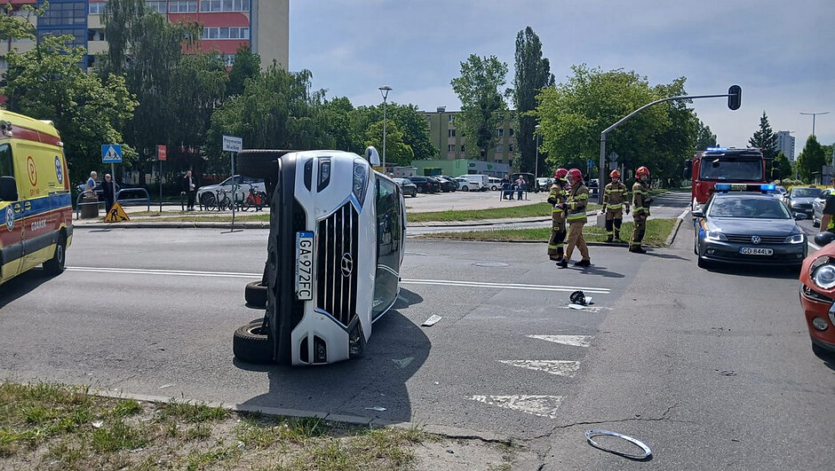 Scena rozgrywa się na ruchliwym skrzyżowaniu w zabudowie miejskiej.
W centrum kadru znajduje się biały Hyundai (GA 972FC) przewrócony na prawy bok. Auto wygląda na świeżo po kolizji — wokół nie ma widocznych śladów ognia, ale sytuacja jest poważna. Wokół pojazdu stoją strażacy w pełnym umundurowaniu, z hełmami i sprzętem ratowniczym. Obok nich znajduje się ambulans, a po prawej stronie radiowóz policyjny z włączonymi światłami. W tle widać również wóz strażacki, co sugeruje, że służby działają w pełnym składzie.
Na chodnikach stoją przechodnie, którzy obserwują akcję ratunkową. W tle widoczne są bloki mieszkalne, drzewa i elementy miejskiej infrastruktury.
