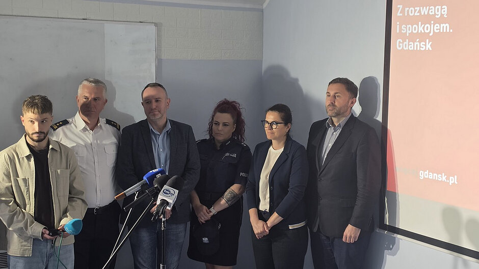 Kontekst i otoczenie
Zdjęcie zostało wykonane w pomieszczeniu przypominającym salę konferencyjną lub szkoleniową. W tle widoczna jest ściana z jasnych bloczków oraz tablica suchościeralna. Po prawej stronie znajduje się ekran projekcyjny z prezentacją.
Osoby i ich ustawienie
Na zdjęciu stoi w rzędzie kilka osób obok siebie. Część z nich jest ubrana w mundury służbowe (widoczne elementy przypominające umundurowanie Straży Miejskiej i Policji), a część w stroje cywilne (marynarki, koszule).
Na pierwszym planie ustawiony jest zestaw mikrofonów na statywie, co wskazuje na oficjalne wystąpienie lub konferencję prasową.
Po lewej stronie jedna z osób trzyma niewielkie urządzenie (prawdopodobnie rejestrator lub mikrofon reporterski).
Tekst widoczny na ekranie
Na ekranie projekcyjnym po prawej stronie widnieje napis:
„Z rozwagą i spokojem. Gdańsk”
oraz adres strony:
„gdansk.pl”
Oświetlenie i szczegóły
oświetlenie jest mieszane: światło naturalne i sztuczne,
część twarzy i sylwetek jest częściowo zacieniona,
kolory są stonowane – dominują biel, granat i szarości.
Charakter sceny
Całość wskazuje na oficjalne spotkanie, briefing lub konferencję dotyczącą działań miejskich lub służb porządkowych.