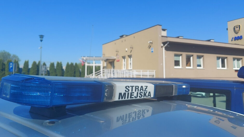 Na pierwszym planie widoczny jest dach pojazdu Straży Miejskiej z niebieskim sygnałem świetlnym i napisem „STRAŻ MIEJSKA”. W tle znajduje się budynek jednostki, z widocznym wejściem głównym oraz godłem na elewacji. Całość przedstawiona jest w słoneczny dzień, na tle bezchmurnego, niebieskiego nieba.