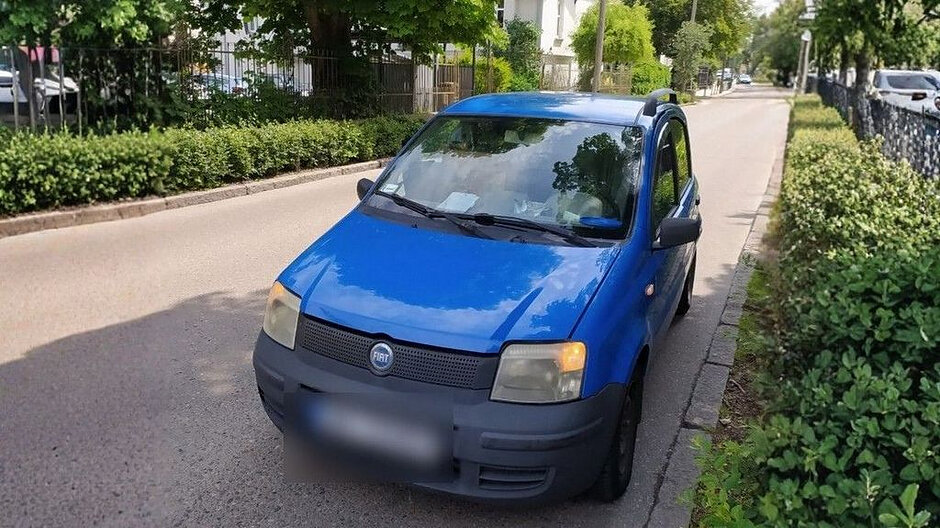 Na zdjęciu widoczny jest niebieski, niewielki samochód osobowy zaparkowany równolegle przy krawężniku na spokojnej, osiedlowej ulicy. Auto stoi po prawej stronie jezdni, częściowo zacienione przez drzewa rosnące wzdłuż chodnika. Wokół znajdują się zadbane krzewy i zielone żywopłoty, oddzielające chodnik od posesji oraz ulicy. W tle po obu stronach drogi widać zaparkowane inne pojazdy oraz budynki mieszkalne o jasnych elewacjach. Nawierzchnia drogi jest równa i czysta, a cała scena sprawia wrażenie spokojnej, dziennej sytuacji w dzielnicy mieszkalnej. Samochód ma widoczne przednie reflektory, grill z logo marki oraz lekko przyciemnioną szybę, a tablica rejestracyjna została zamazana.