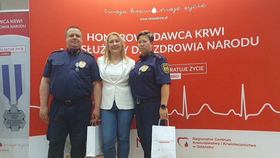 Na zdjęciu przedstawiono trzy osoby stojące obok siebie na tle czerwonej ścianki promocyjnej związanej z krwiodawstwem. Tło zawiera liczne napisy, w tym widoczny, duży tekst „HONOROWY DAWCA KRWI ZASŁUŻONY DLA ZDROWIA NARODU”, a także hasło „twoja krew – moje życie” oraz nazwę strony internetowej i odniesienie do Regionalnego Centrum Krwiodawstwa i Krwiolecznictwa w Gdańsku. Na ściance widoczny jest również motyw graficzny przypominający zapis EKG oraz elementy związane z symboliką krwi i ratowania życia. Dwie osoby stojące po bokach ubrane są w ciemne uniformy służbowe z naszywkami i odznaczeniami przypiętymi do piersi. W ich rękach widoczne są białe torby. Pomiędzy nimi stoi osoba ubrana w jasną marynarkę i spodnie, również trzymająca torbę. Całość sprawia wrażenie oficjalnego wydarzenia lub uroczystości związanej z wyróżnieniem osób zaangażowanych w działalność krwiodawczą. Kolorystyka zdjęcia zdominowana jest przez intensywną czerwień tła, kontrastującą z ciemnymi mundurami i jasnym strojem osoby stojącej pośrodku.