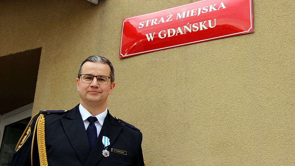 Na zdjęciu widoczna jest osoba stojąca na zewnątrz budynku o jasnej, beżowej elewacji. W górnej części kadru znajduje się czerwona tablica z białym napisem „STRAŻ MIEJSKA W GDAŃSKU”, przymocowana do ściany. Na pierwszym planie, po lewej stronie obrazu, znajduje się osoba ubrana w ciemny, formalny mundur z wyraźnymi elementami ozdobnymi, takimi jak pagony, złoty sznur naramienny oraz odznaka przypięta do piersi. Na mundurze widoczny jest także medal zawieszony na wstążce oraz niewielka tabliczka z napisem. Osoba stoi przodem do obiektywu, a za nią, po lewej stronie kadru, częściowo widoczne jest wejście do budynku lub wnęka drzwiowa. Tło stanowi jednolita, chropowata powierzchnia ściany, co sprawia, że główna postać i czerwona tablica informacyjna są najbardziej wyeksponowanymi elementami fotografii. Kolorystyka zdjęcia jest stonowana, z dominacją beżu i czerni, przełamanych intensywną czerwienią tablicy.