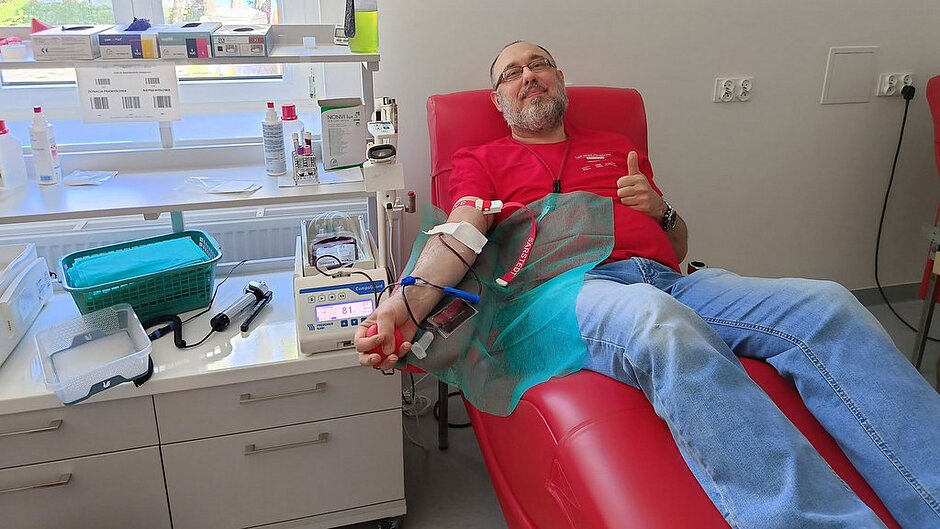 Na zdjęciu przedstawiono wnętrze pomieszczenia medycznego przeznaczonego do oddawania krwi. W centralnej części kadru znajduje się osoba leżąca na czerwonym, regulowanym fotelu medycznym. Ramię tej osoby jest podłączone do aparatury do poboru krwi za pomocą przewodów i igły, a w dłoni trzyma ona gumową piłeczkę używaną podczas procedury. Na przedramieniu widoczna jest opaska uciskowa oraz zabezpieczenia medyczne. Obok fotela, po lewej stronie, ustawiony jest sprzęt medyczny z wyświetlaczem oraz pojemnik na pobieraną krew. Na blacie i półkach znajdują się różne akcesoria medyczne, takie jak rękawiczki, pojemniki, środki dezynfekujące, probówki oraz koszyki na materiały jednorazowe. W tle widoczne jest okno, przez które wpada naturalne światło, a za szybą można dostrzec drzewa i fragment budynku. Ściany pomieszczenia są jasne i jednolite, co podkreśla sterylny charakter miejsca. Całość przedstawia moment oddawania krwi w warunkach medycznych, z wyraźnie widoczną aparaturą oraz przygotowanym stanowiskiem do przeprowadzania tego typu procedur.